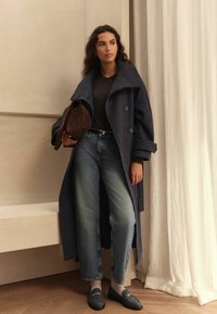 Long navy checkered coat with large collar, black t-shirt, loose jeans, gray socks, and dark loafers. Brown bag in hand, standing near curtains.