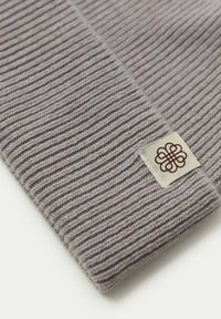 Grå ribbad mössa i mjukt material, med en krämfärgad etikett med en brun blommig logotyp. Den strukturerade designen bidrar med visuell intressanthet.