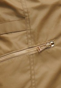 Οριζόντια χρυσή φερμουάρ σε ταμπα ύφασμα με ορατές ραφές, ελαφρώς ανοιχτή με το τσεπώ στη δεξιά πλευρά.