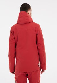 Rote wasserdichte Jacke mit Kapuze, ausgestattet mit einer glatten Textur, Nahtdetails und einem Logotag an der Seite. Rückansicht dargestellt.