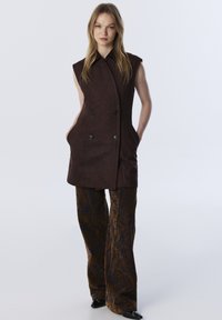 Brauner ärmelloser Mantel mit doppelreihigem Design, ausgestattet mit schwarzen Knöpfen und Seitentaschen, kombiniert mit ausgestellten gemusterten Hosen.