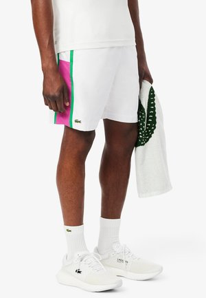 Białe spodenki z różowymi i zielonymi paskami po bokach, z zielonym logo Lacoste. Model nosi białe sneakersy i trzyma białą ręcznik.