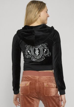 Žena nosi crnu velurnu jaknu s kapuljačom s bijelim Juicy Couture grbom na leđima i smeđe velurne hlače s džepovima na stražnjoj strani.