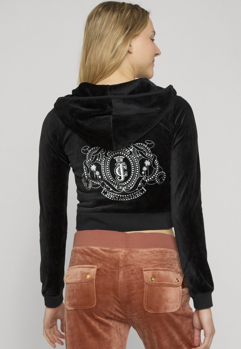 Femme portant une veste en velours noire à capuche avec un logo blanc Juicy Couture sur le dos et un pantalon en velours marron avec des poches arrière.