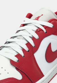 Close-up do sneaker Nike Air Jordan branco e vermelho, mostrando os atacadores brancos, o swoosh vermelho e o logo Jumpman na língua.