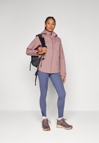 Ljusrosa vattentät jacka med huva, i kombination med blå leggings och bruna vandringsskor. Modellen håller en liten svart ryggsäck.