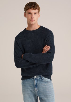 Suéter de punto azul marino con textura de canalé y corte relajado. Presenta un escote redondo y mangas largas, combinado con jeans de denim claro.