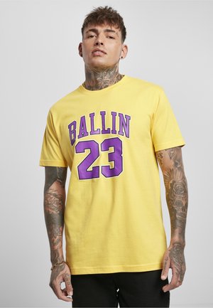Tetoválásokkal rendelkező férfi sárga pólót visel lila szöveggel: "BALLIN 23", egy egyszínű háttér előtt áll.