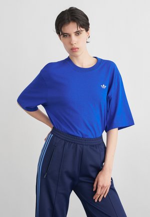 Ung person med kort mørkt hår iført en blå Adidas T-shirt og marineblå træningsbukser med blå striber, stående foran en ensartet lys baggrund.
