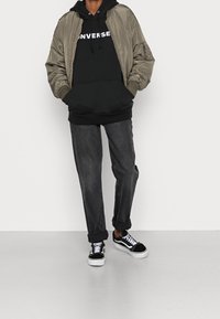 Giubbotto bomber oliva, felpa nera con testo bianco, jeans scuri, sneaker bianche e nere. I materiali sembrano lisci e informali.