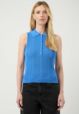Kvinde med langt blond hår iført blå ærmeløs ribbet poloshirt og sorte jeans, stående mod en ensfarvet lys baggrund.