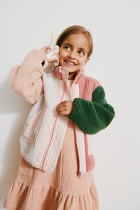 Μπουφάν από φλις με πολλαπλά χρώματα που περιλαμβάνει ροζ, πράσινο και λευκά πάνελ, με απαλή υφή και επένδυση με κεντήματα· φορεμένο πάνω από ένα ριμπ ροζ φόρεμα.