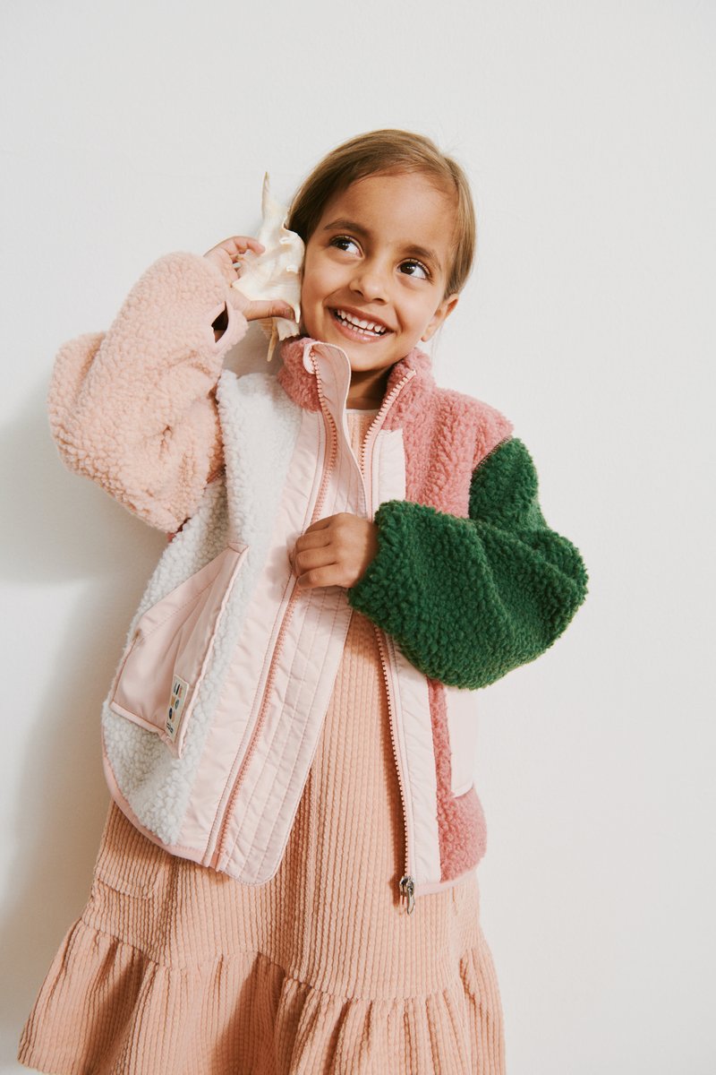 Μπουφάν από φλις με πολλαπλά χρώματα που περιλαμβάνει ροζ, πράσινο και λευκά πάνελ, με απαλή υφή και επένδυση με κεντήματα· φορεμένο πάνω από ένα ριμπ ροζ φόρεμα.