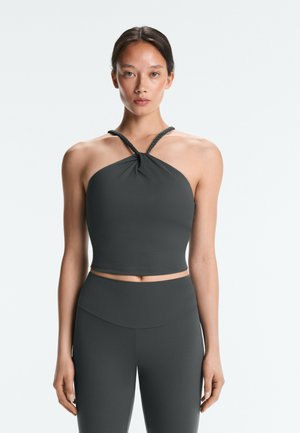 Jeune femme debout, face à l’avant, portant un crop top sans manches gris foncé style halter et un legging taille haute assorti, sur un fond uni.