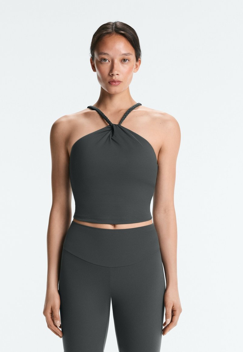 Jeune femme debout, face à l’avant, portant un crop top sans manches gris foncé style halter et un legging taille haute assorti, sur un fond uni.