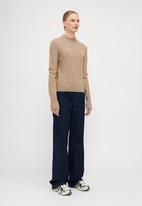 Beige strikkegenser med høy hals og ribbestruktur, kombinert med mørkeblå vide jeans og hvite joggesko med svarte detaljer.