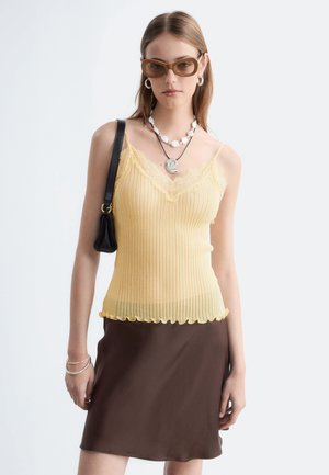 Jeune femme portant des lunettes de soleil marron, un débardeur jaune à dentelle, une jupe marron et un sac à bandoulière noir sur fond blanc.