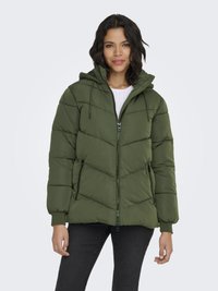 Woman wearing a green puffer jacket with zipper pockets, hood, and black pants, standing against a plain light background.