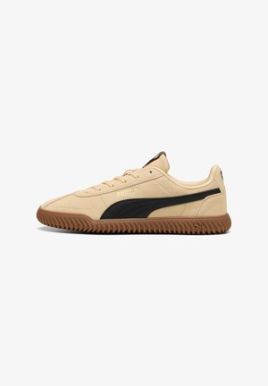 Zapatillas Puma con parte superior de ante beige, franja lateral negra y suela de goma marrón estriada. Cuenta con diseño de cordones y cuello acolchado.