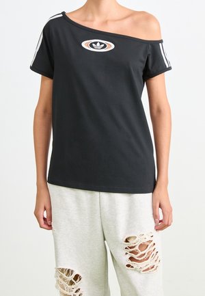 Personne portant un t-shirt Adidas noir à épaules dénudées avec des bandes blanches sur les manches et un pantalon gris clair avec de grandes déchirures aux cuisses.
