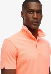 Polohemd in fel koraal; korte mouwen, zachte katoenen stof, platte kraag met drieknoppenlijst, merketiket op de mouw.