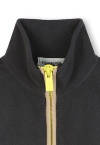 Dunkelgraue Fleecejacke mit hohem Kragen, ausgestattet mit einem gelben Reißverschluss und beigen Akzenten entlang des Reißverschlusses. Etikett im Kragen sichtbar.