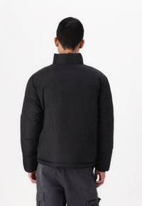 Jachetă puf din material negru, cu un guler înalt, croială ușor supradimensionată și detalii de cusătură vizibile. Partea din spate prezintă un design subtil cu cusături în formă de V.