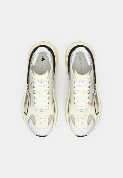 Para białych i szarych sportowych sneakersów z sznurówkami, czarnymi i beżowymi akcentami oraz czerwonym detalem z logo, pokazanych z góry na białym tle.