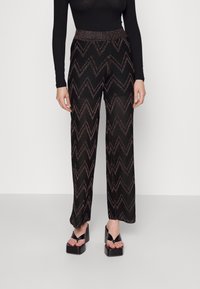 Kobieta ubrana w czarne spodnie o szerokich nogawkach z zygzakowatym wzorem, czarną bluzkę z długim rękawem oraz czarne sandały na platformie z odkrytymi palcami.