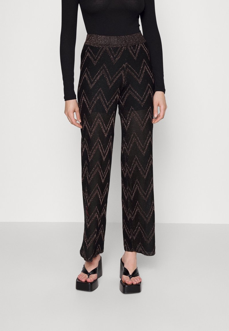 Kobieta ubrana w czarne spodnie o szerokich nogawkach z zygzakowatym wzorem, czarną bluzkę z długim rękawem oraz czarne sandały na platformie z odkrytymi palcami.