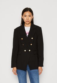 Mustat kaksiriviset bleiserit, jotka on valmistettu sileästä kankaasta, jossa on kultaiset napit, leikattu kaulus ja sivutaskut yhdistettynä sinisiin farkkuihin.