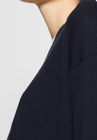 Gros plan sur le cou et l'épaule d'une personne portant un pull en tricot bleu marine foncé sur un fond clair uni.