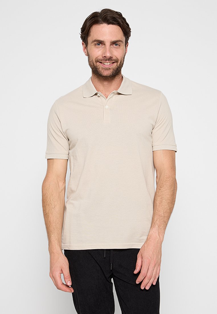 Polo à manches courtes beige en mélange de coton, avec un plastron à deux boutons et un col classique. Associé à un pantalon noir.