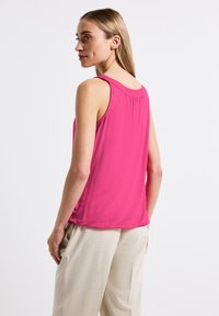 Top senza maniche fucsia con scollatura arricciata, texture liscia e design fluido, abbinato a pantaloni beige chiaro, dalla vestibilità comoda.