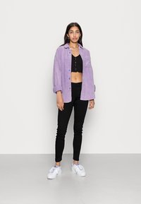 Camisa de pana morada, top negro corto, jeans negros ajustados y zapatillas blancas. La camisa tiene un frente abotonado y mangas enrolladas.