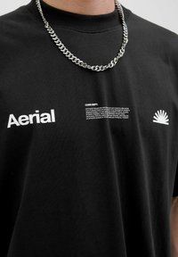 Camiseta de algodón negra con la palabra "Aerial" en texto blanco, con un gráfico en el pecho derecho y un collar de cadena plateada puesto sobre ella.