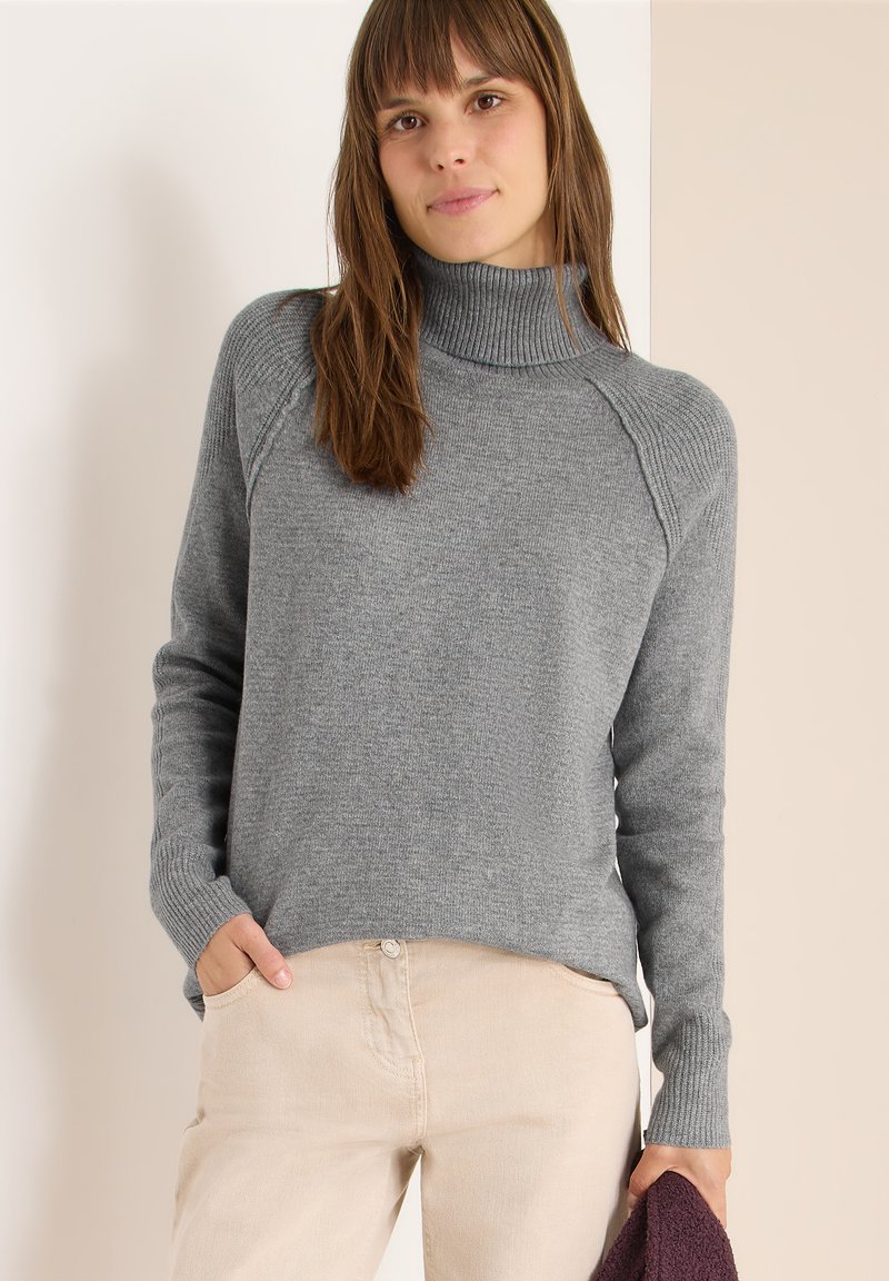 Pull-over en col roulé gris avec des poignets et un ourlet côtelés, présentant une coupe décontractée et un tissu légèrement texturé. Associé à un pantalon beige clair.