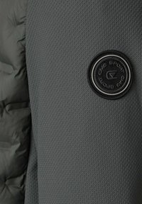 Mörkgrå texturerad tyg med en rund svart "CMS SPORT" logotyp patch sydd på högra sidan, bredvid slätt vadderat material.