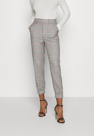 Femme portant un pantalon à carreaux taille haute avec des revers, associé à un haut blanc à manches longues et des talons noirs à bride cheville, mains dans les poches.