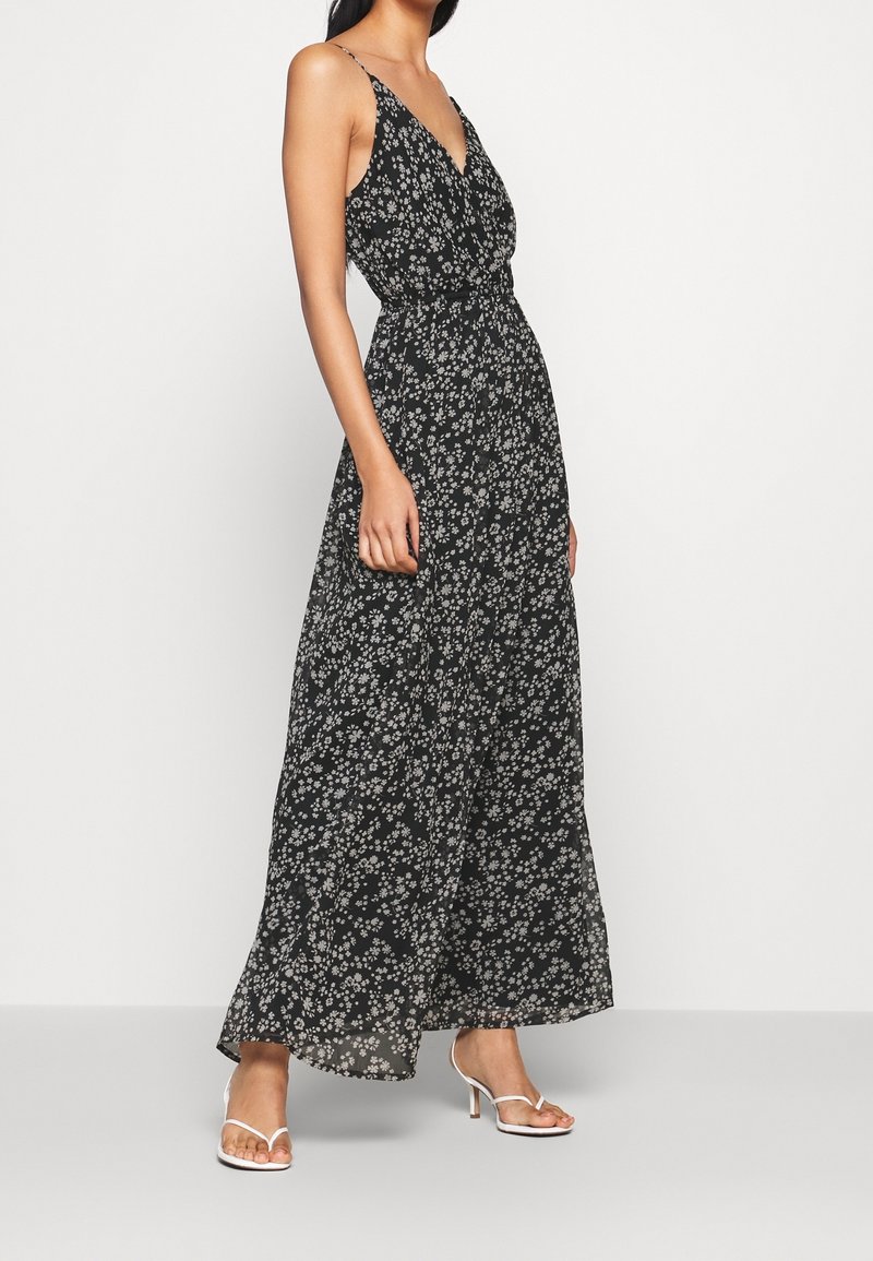 Femme portant une robe longue noire avec un imprimé floral blanc, des bretelles spaghetti et un décolleté en V, associée à des sandales blanches à talons.