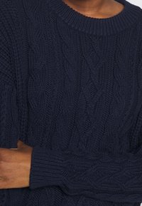 Laivastonsininen palmikkoneulepusero, jossa on pyöreä pääntie ja resorit hihansuissa, esittää monimutkaisia punottuja kuvioita koko kankaan läpi.