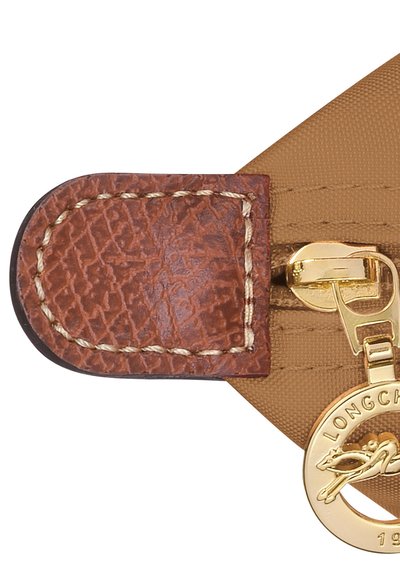 Brown leather accent with textured finish, gold zipper pull, and logo charm attached to tan fabric surface. Stitched detailing visible.