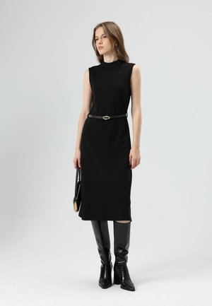Zwarte mouwloze jurk met een aansluitend ontwerp, knielengte, gedragen met een bruine riem met een zilveren gesp, gecombineerd met kniehoge zwarte laarzen.
