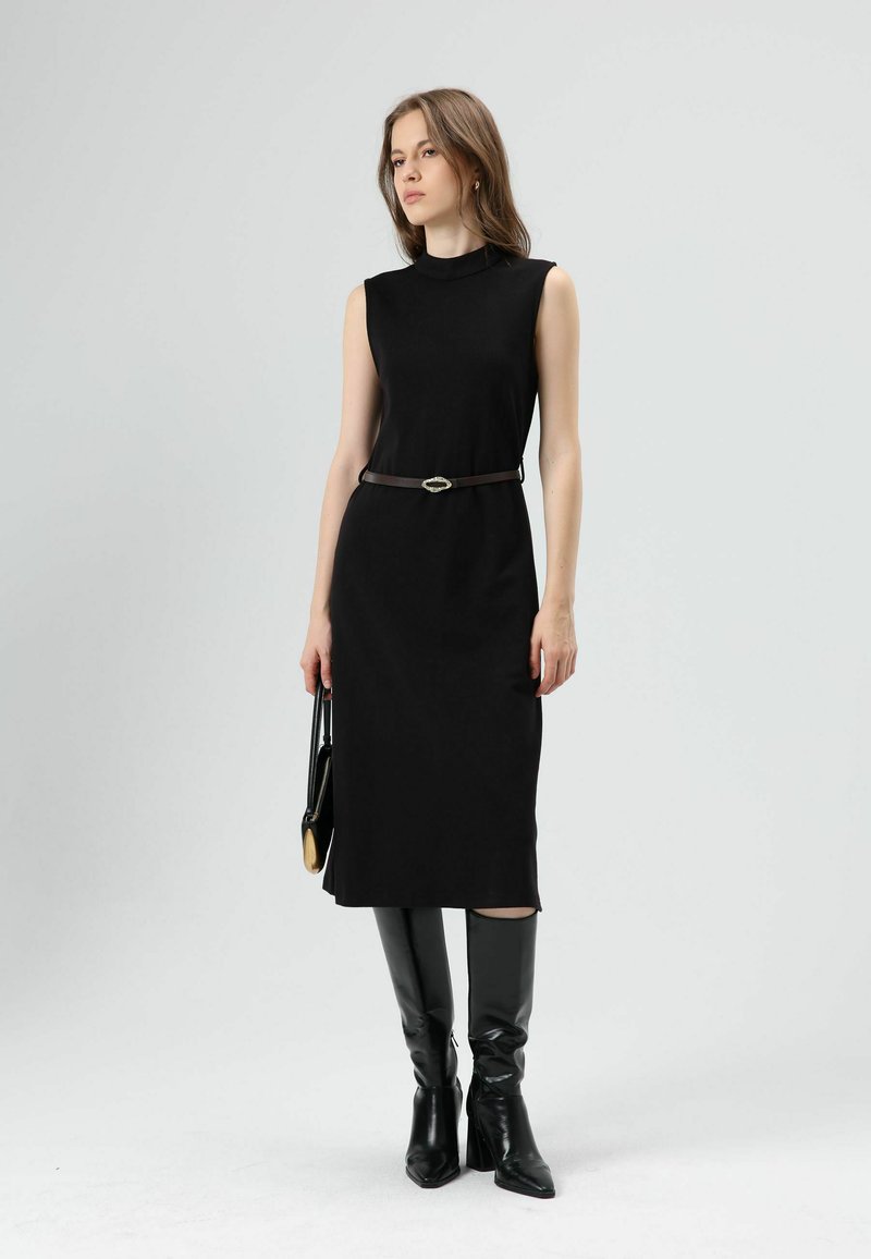 Zwarte mouwloze jurk met een aansluitend ontwerp, knielengte, gedragen met een bruine riem met een zilveren gesp, gecombineerd met kniehoge zwarte laarzen.