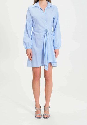 Femme portant une robe bleu clair à manches longues, boutonnée, avec un détail noué à l'avant et des sandales argentées à talons hauts et fines lanières, debout sur fond blanc.