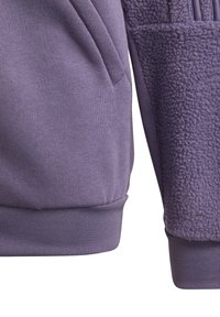 Lila hoodie med en mjuk, texturerad fleecepanel; har ribbade muddar och fåll, samt en sidficka på den släta tygdelen.