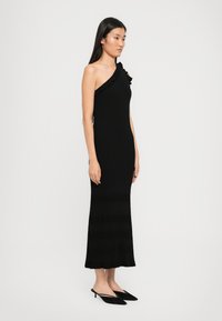 Nő áll egy fekete, egyvállas maxi ruhában, fodros részletekkel, fekete hegyes orrú slingback kisméretű magassarkú cipőben, egy sima háttér előtt.