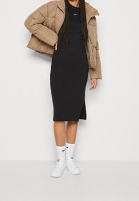 Puffjacka i beige, svart tajt klänning, vita strumpor och vita sneakers. Jackan har en hög krage och dragkedjestängning. Klänningen har en knähög slits.