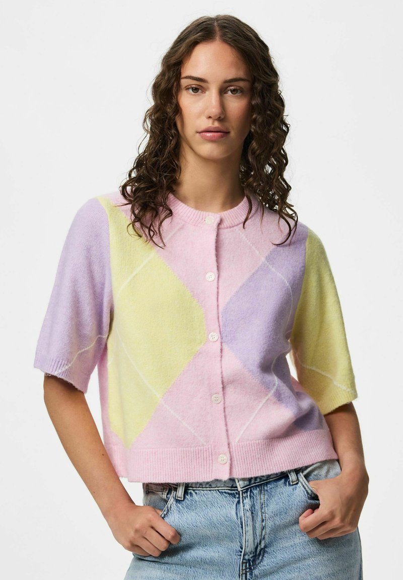 Femme aux cheveux bouclés portant un cardigan argyle pastel à manches courtes et un jean bleu clair, debout les mains dans les poches sur fond blanc.