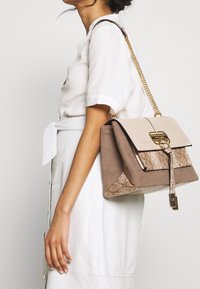 Bolso en tonos marrón y crema con cierre de solapa, herrajes dorados y asa de cadena. Presenta detalles de textura de piel de serpiente y una forma estructurada.